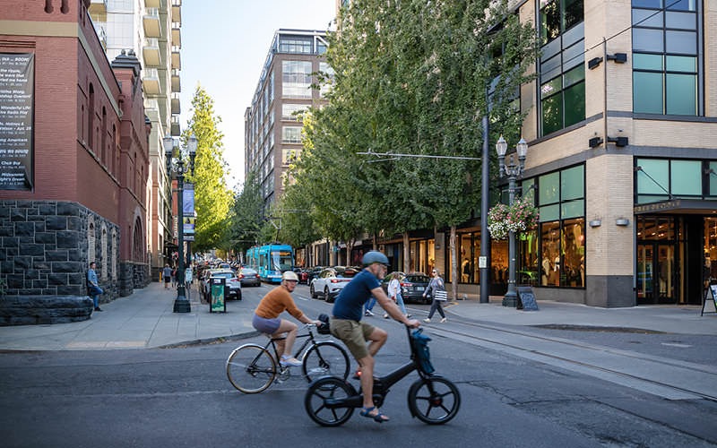 men ride bicycles through downtown as people cross city street